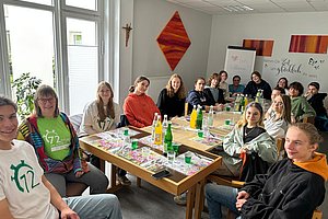 Gruppe von Jugendlichen sitzt an zwei zusammengestellten Tischen in einem Raum mit Wanddekoration und Getränken auf den Tischen