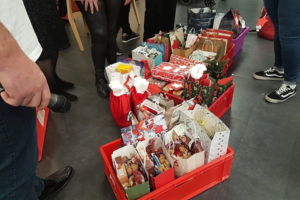 Mehrere rote Kisten mit Geschenkverpackungen und Geschenktüten auf dem Boden, umgeben von Personen, von denen nur Beine und Füße sichtbar sind