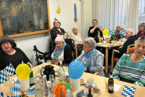 Gruppe älterer Menschen sitzt an Tischen mit blau-weißen Papierfahnen, Luftballons und Getränken in einem hellen Raum