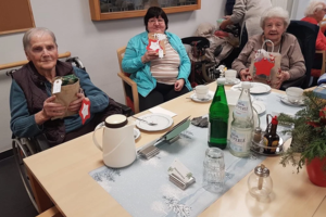 Mehrere ältere Menschen sitzen an einem Tisch mit Tassen, Flaschen und einem Weihnachtsgesteck und halten Geschenke in den Händen