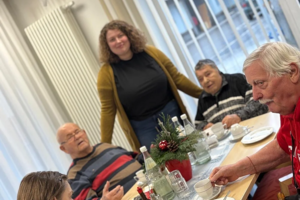 Mehrere Personen sitzen an einem Tisch mit Tassen, Flaschen und einem kleinen Weihnachtsgesteck in einem hellen Raum mit Fenster und Heizkörper.