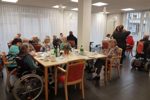Mehrere ältere Menschen sitzen an zwei Tischen in einem hellen Raum, einige im Rollstuhl, andere auf Stühlen, mit Getränken und Geschirr auf den Tischen.
