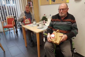 Zwei ältere Männer in einem Raum mit Tisch und Stühlen, einer sitzt im Rollstuhl und hält ein Geschenk, der andere sitzt am Tisch und hält eine Tasche mit Weihnachtsmotiv