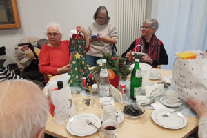 Mehrere ältere Personen sitzen an einem Tisch mit Tassen, Tellern, Getränkeflaschen und weihnachtlichen Geschenktüten