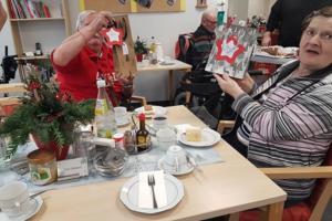 Zwei Personen sitzen an einem Tisch mit Kaffeetassen und Kuchen und halten jeweils eine Geschenkverpackung mit rotem Stern hoch