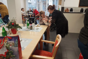 Pflegekraft spricht mit älterer Frau im Rollstuhl in einem Gemeinschaftsraum mit mehreren älteren Menschen und weihnachtlicher Dekoration auf dem Tisch