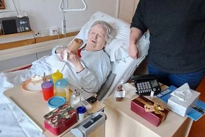 Krankenbett mit älterer Person, die ein Getränk hält, daneben ein Nachttisch mit Medikamenten und Pflegeutensilien, im Hintergrund ein Mann stehend