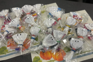 Mehrere kleine Gespenster aus weißem Stoff mit schwarzen Augen, die in durchsichtigen Folienbeuteln mit bunten Bändern verpackt sind, liegen auf einer Tischdecke mit Kürbismotiv
