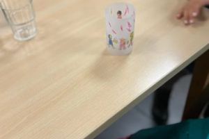 Holztisch mit einem Glas Wasser und einem kleinen, bemalten Papierbecher, zwei Personen sitzen am Tisch, von denen nur Hände und Teile der Kleidung sichtbar sind