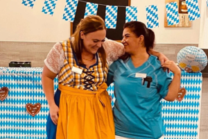 Zwei Frauen stehen nebeneinander, eine trägt ein traditionelles Dirndl mit gelbem Rock, die andere eine blaue Arbeitskleidung, im Hintergrund hängen blau-weiße Rautenfahnen und Dekorationen mit Lebkuchenherzen.
