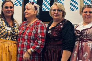 Vier Frauen in traditioneller bayerischer Tracht stehen nebeneinander vor einer Wand mit einer Girlande aus blau-weißen Rauten.