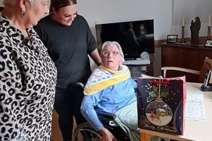 Drei ältere Personen in einem Wohnzimmer, eine sitzt auf einem Stuhl mit einem Buch auf dem Schoß, zwei stehen daneben.