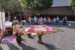 Gruppe älterer Menschen, einige im Rollstuhl, sitzen im Halbkreis auf einem gepflasterten Platz vor einem großen, bunten Blumenmuster auf dem Boden