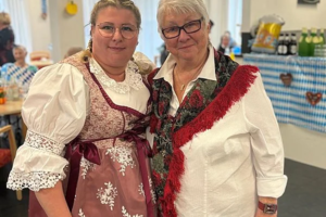 Zwei Frauen in traditioneller Tracht stehen in einem festlich dekorierten Raum mit blau-weißen Girlanden und Tischen im Hintergrund
