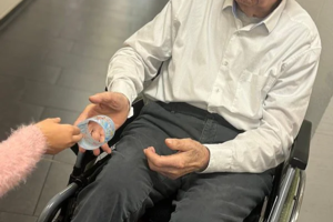 Holztisch mit einem Glas Wasser und einem kleinen, bemalten Papierbecher, zwei Personen sitzen am Tisch, von denen nur Hände und Teile der Kleidung sichtbar sind
