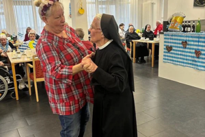 Zwei Frauen tanzen in einem festlich geschmückten Raum mit mehreren sitzenden Gästen im Hintergrund