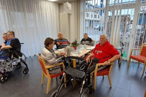 Mehrere ältere Menschen sitzen in einem Gemeinschaftsraum an Tischen und unterhalten sich teilweise