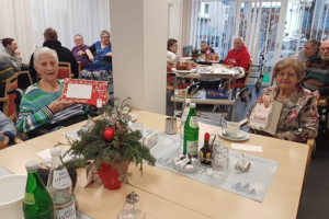 Mehrere ältere Menschen sitzen an Tischen in einem hellen Raum, auf einem Tisch steht eine Weihnachtsdekoration mit Tannenzweigen und roten Kugeln