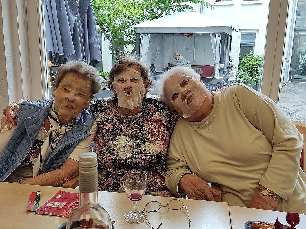 Drei ältere Personen sitzen eng nebeneinander an einem Tisch mit Weinflasche, Weinglas und Brille, alle drei tragen eine Gesichtsmaske.