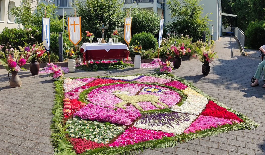 Blumenbild mit einem Lamm auf einem roten und weißen Hintergrund auf einem gepflasterten Platz vor einem Altar mit drei Fahnen und Blumengestecken