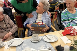 Mehrere ältere Menschen sitzen an einem gedeckten Tisch, eine Person in grüner Kleidung überreicht einer sitzenden Person ein Geschenk.