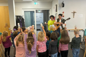 Kinder stehen in einem Raum und halten Becher nach oben, während zwei Erwachsene vorne stehen, einer mit einer Gitarre.