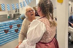 Zwei Personen halten sich an den Händen, eine trägt ein traditionelles Dirndl mit geflochtenem Haar, im Hintergrund blau-weiß karierte Dekoration und Lebkuchenherzen an der Wand