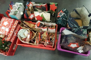 Mehrere bunte Geschenktüten und -verpackungen mit Weihnachtsmotiven in roten und lila Kunststoffkisten auf grauem Boden