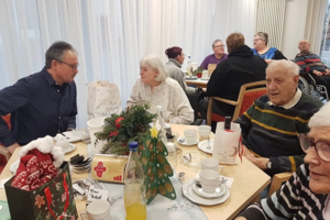 Mehrere ältere Menschen sitzen an einem Tisch mit Weihnachtsdekoration und Geschirr in einem hellen Raum.