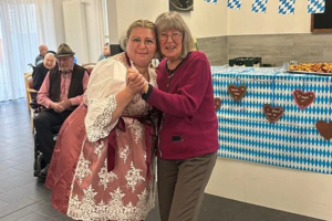 Zwei Frauen tanzen in einem Raum mit blau-weiß karierten Girlanden und einer Theke mit Lebkuchenherzen im Hintergrund