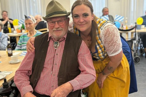 Mann im Rollstuhl mit Hut und Weste neben Frau in traditioneller Tracht mit gelbem Rock in einem festlich dekorierten Raum mit mehreren Personen im Hintergrund