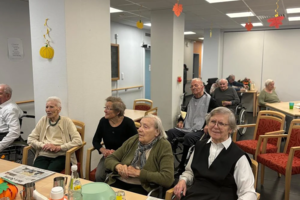 Mehrere ältere Menschen sitzen in einem Gemeinschaftsraum an Tischen mit Zeitungen und Getränken, im Hintergrund sind herbstliche Dekorationen an der Decke zu sehen.